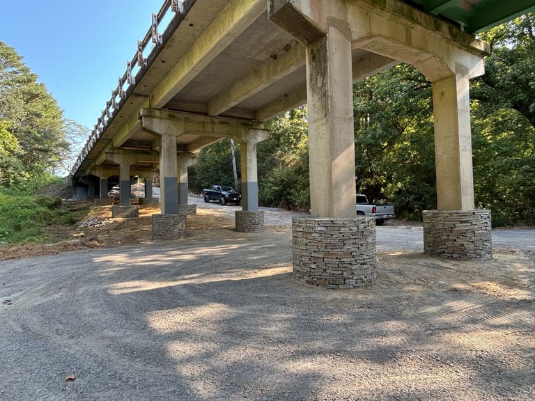 Concrete underpass with large support columns and stone-faced pillars, surrounded by green trees and parked cars
