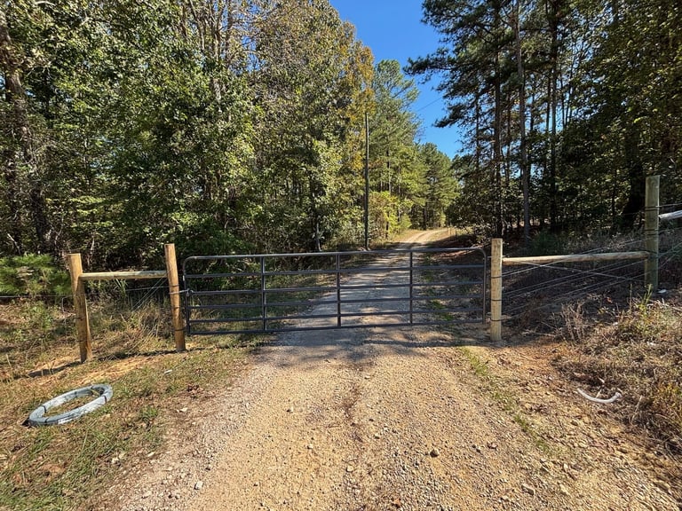 Metal gate across dirt driveway entrance with wooden fence posts and forest trees