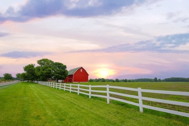 Red barn with white fence in green pasture at sunset with trees and cloudy sky